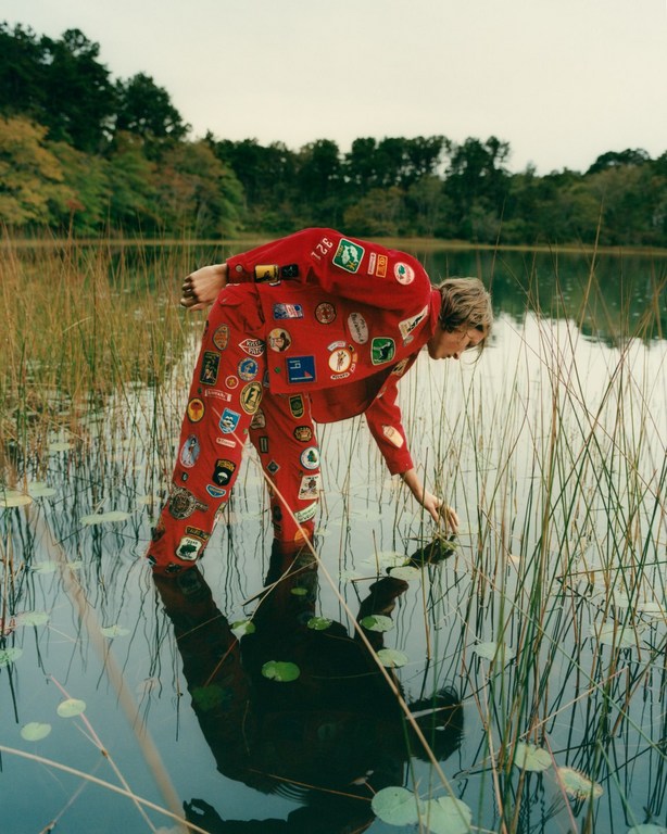 Person in red patch-covered suit stands in shallow lake water near reeds and trees, reaching into the water. Reflection visible.