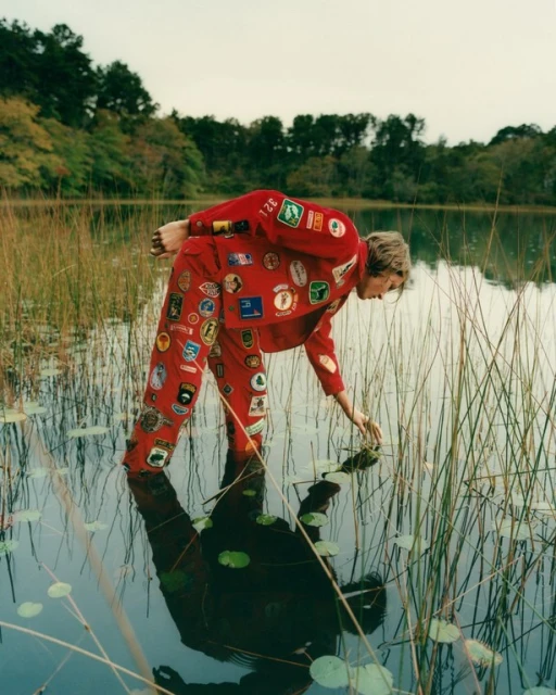 Person in red patch-covered suit stands in shallow lake water near reeds and trees, reaching into the water. Reflection visible.