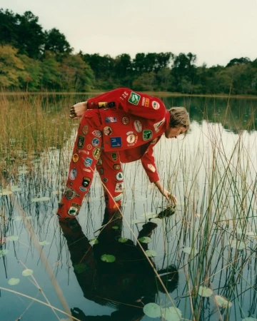 Person in red patch-covered suit stands in shallow lake water near reeds and trees, reaching into the water. Reflection visible.
