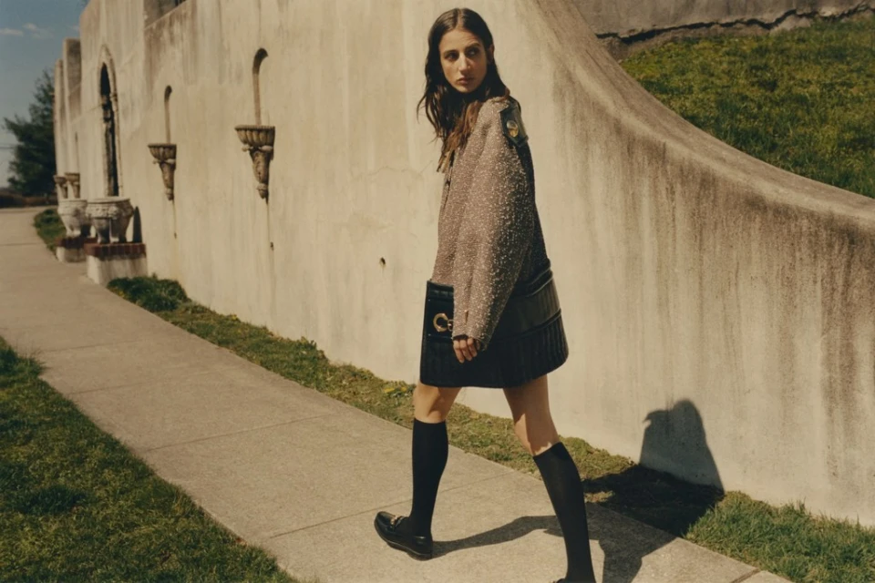 An adult female strides along a concrete walkway beside a light stucco wall. She wears a tweed jacket, dark skirt, and knee-high socks.