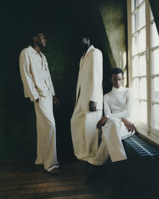 Three dark-skinned men pose in cream-colored outfits against a dark green wall and window. Fashion editorial photography indoors.