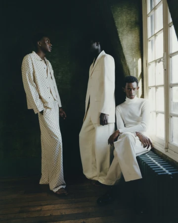 Three dark-skinned men pose in cream-colored outfits against a dark green wall and window. Fashion editorial photography indoors.