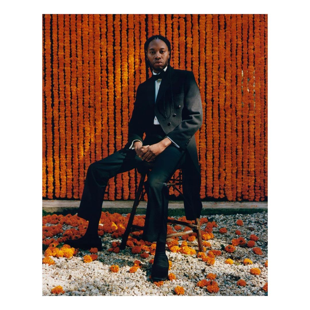 Man in a tuxedo sits on a wooden stool amid a scattering of orange flowers, with a backdrop of dense orange flower garlands.
