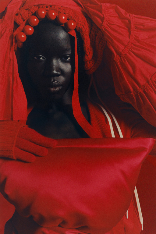 Striking portrait of a dark-skinned woman in a red headpiece with decorative balls, complemented by a matching red top with white stripes.