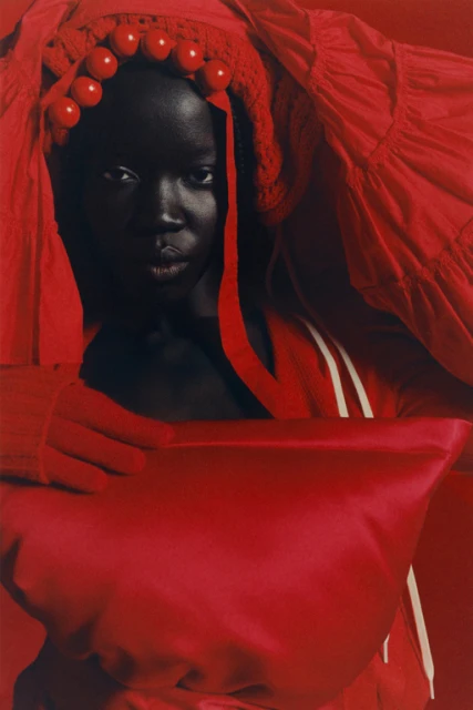 Striking portrait of a dark-skinned woman in a red headpiece with decorative balls, complemented by a matching red top with white stripes.