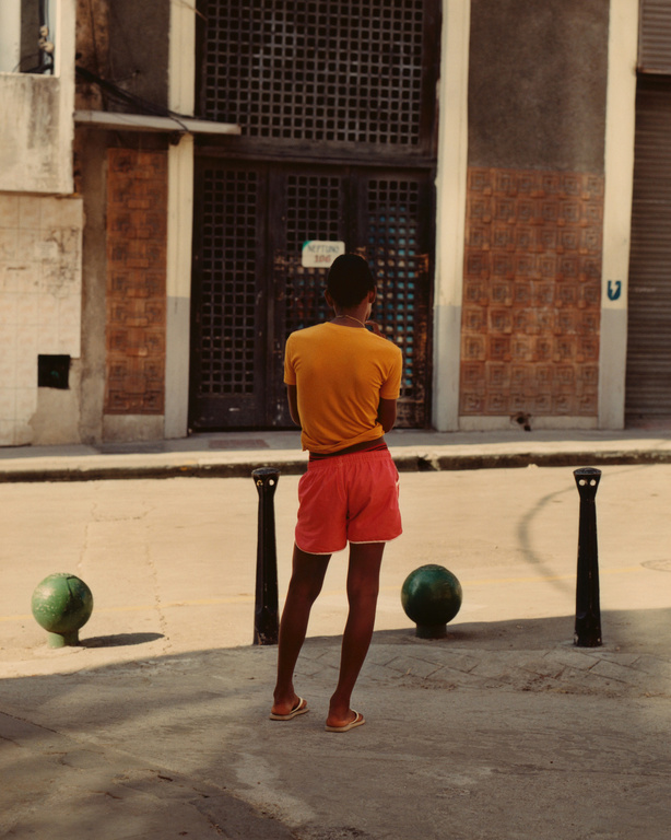 A person stands in a sunny urban setting wearing an orange t-shirt and red shorts, gazing at a building with a lattice door.