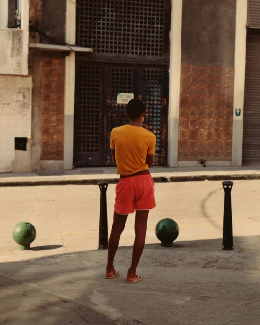 A person stands in a sunny urban setting wearing an orange t-shirt and red shorts, gazing at a building with a lattice door.