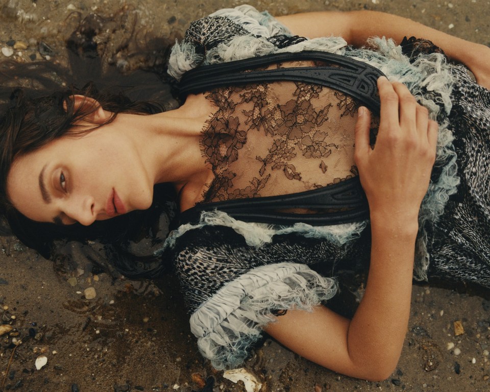 Brunette woman lies on the sandy beach in a lace and tulle dress, looking off to the side in a fashion editorial style portrait.