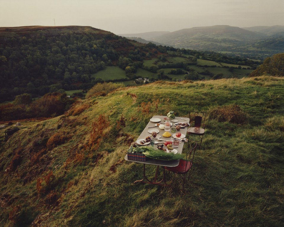 Outdoor dining scene on grassy hilltop with a set table featuring plates, glasses, food, and chairs overlooking a green mountain landscape.