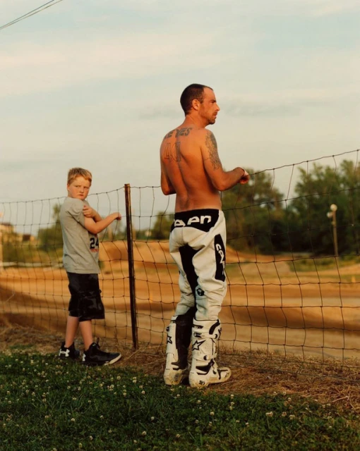 A shirtless man and a young boy stand by a dirt bike track fence on a sunny day, wearing moto attire and looking toward the track.
