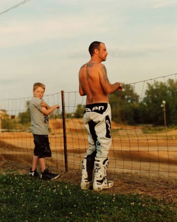A shirtless man and a young boy stand by a dirt bike track fence on a sunny day, wearing moto attire and looking toward the track.