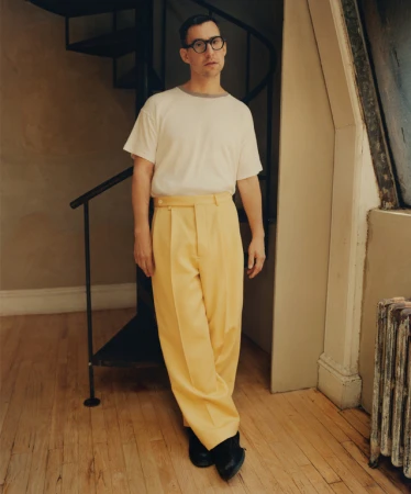 Man wearing round glasses, white t-shirt, and yellow trousers standing in front of a spiral staircase.
