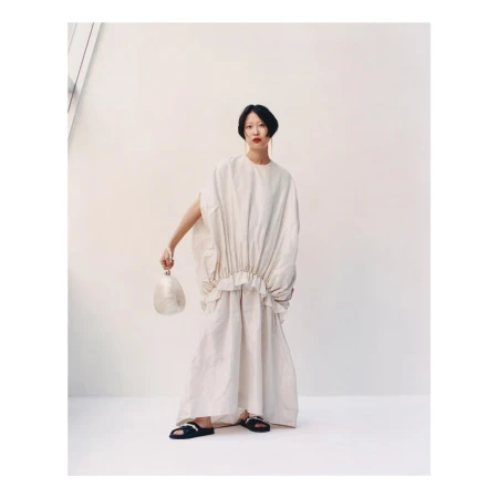 Model in voluminous cream tunic dress holding a small spherical bag against a plain white studio wall.