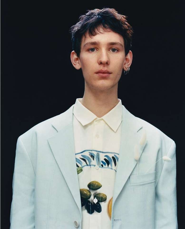 Portrait of a young man wearing a light blue blazer over a patterned white button-down shirt against a dark background.