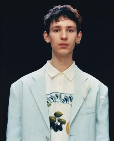 Portrait of a young man wearing a light blue blazer over a patterned white button-down shirt against a dark background.