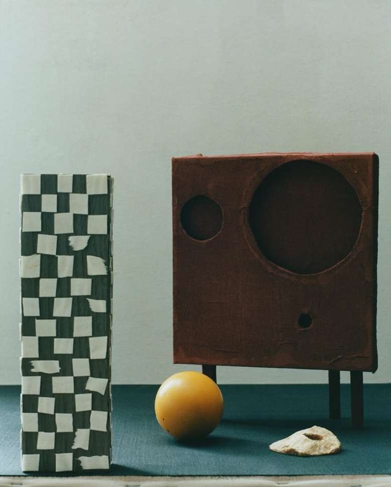 Still life composition featuring a checkered box, a brown cube with circular cutouts, an orange sphere, and a piece of dried fruit.