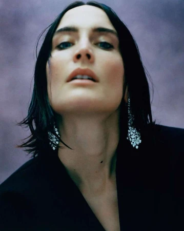 Low-angle portrait of a woman with dark hair wearing sparkling drop earrings and a black jacket against a muted textured background.