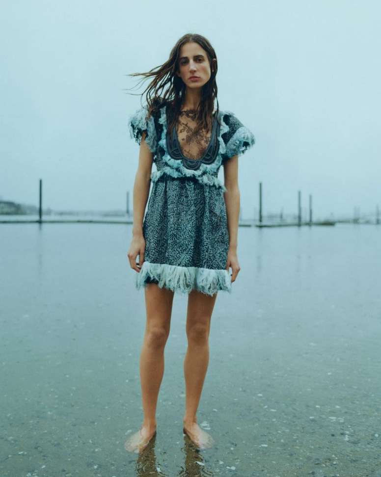 Model wearing a patterned short dress with feathered trim standing in shallow water outdoors by wooden posts