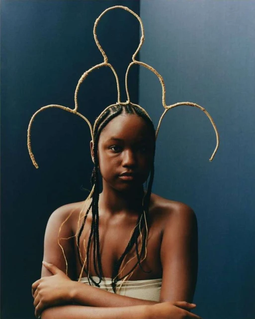 Close-up portrait of a young adult with gold headdress, braids, light wrap, and neutral expression against a blue wall backdrop in a photo studio.