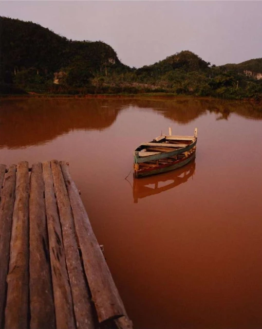Tranquil scene of a wooden boat floating on orange water with lush, green mountains in the background under a muted sky.