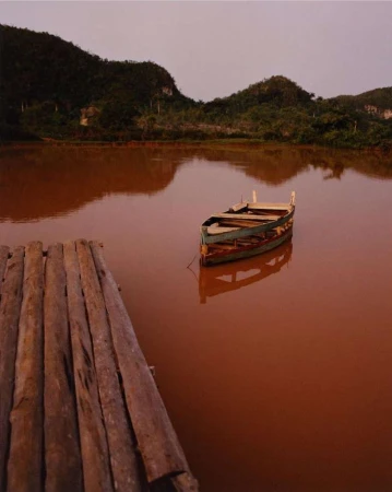 Tranquil scene of a wooden boat floating on orange water with lush, green mountains in the background under a muted sky.
