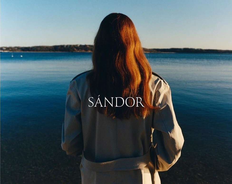 A woman with long red hair, seen from the back, wears a beige trench coat and gazes out over a blue body of water in natural light.