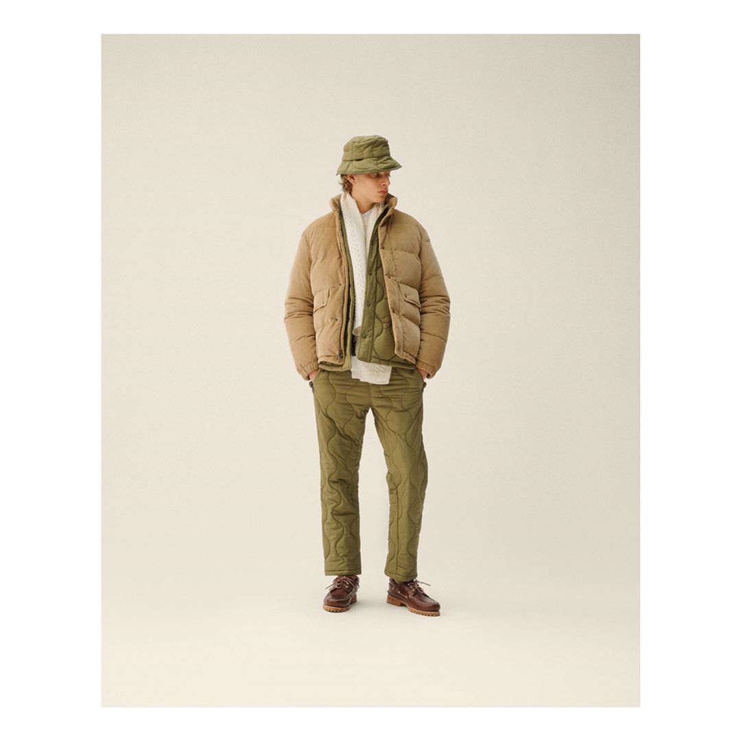Man in a tan puffer jacket, quilted vest and pants, and green bucket hat posing indoors in a fashion editorial photograph.