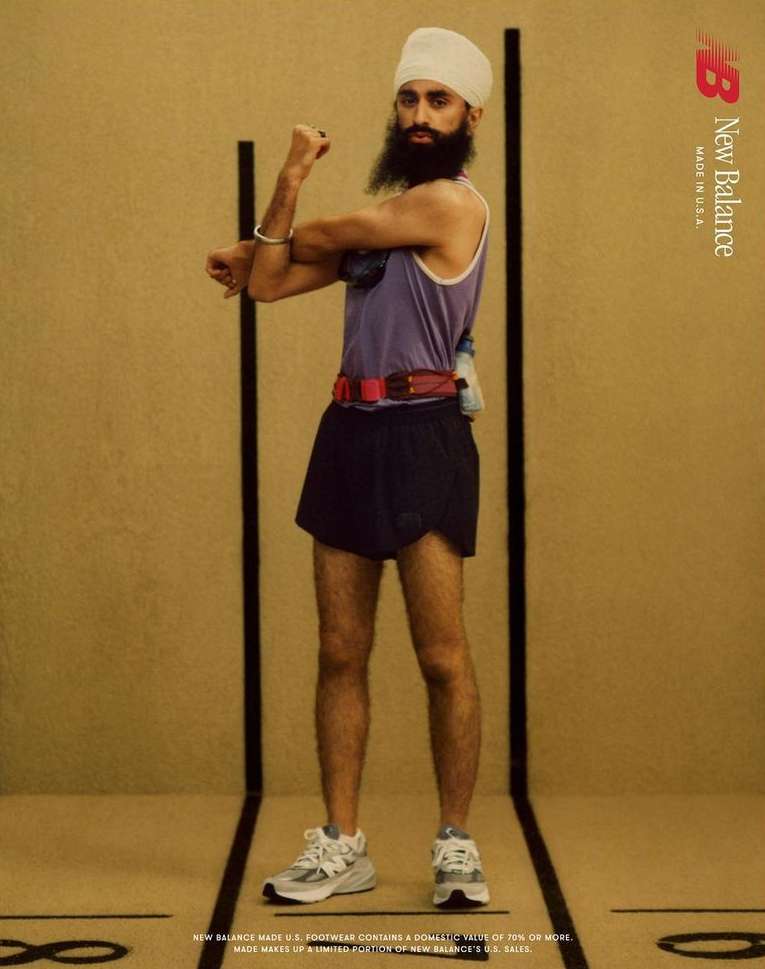 An adult male wearing athletic shorts, a tank top, and New Balance running shoes poses in a studio with a tan background for an advertisement.