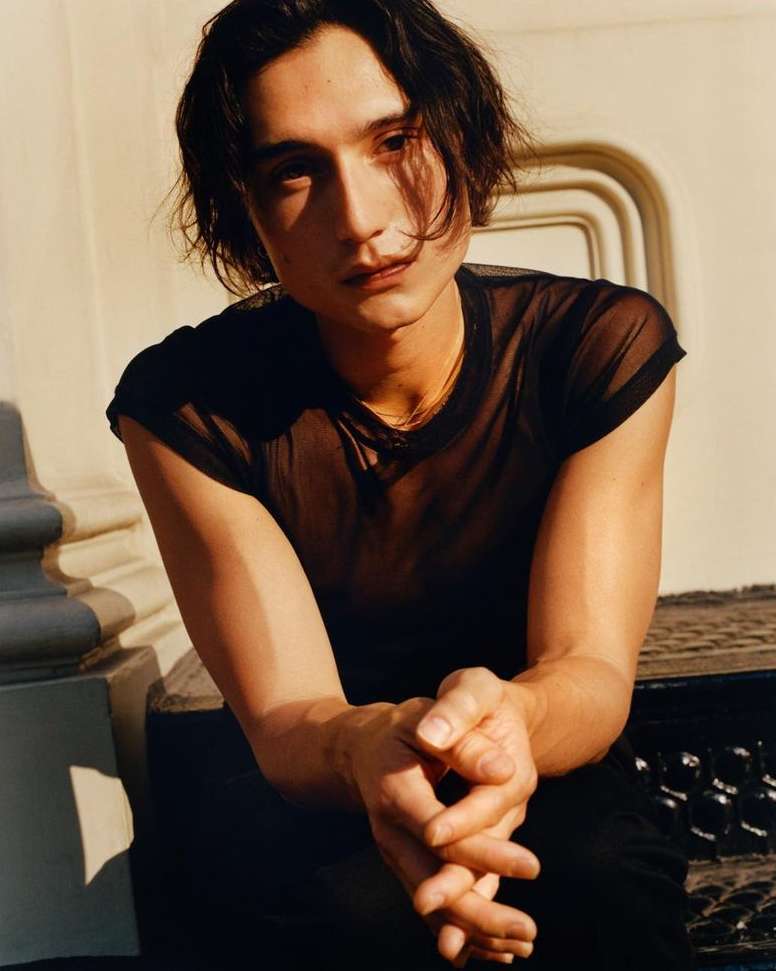 Portrait of a young adult male with dark hair and a sheer black shirt sitting outdoors against a light-colored concrete wall and steps.