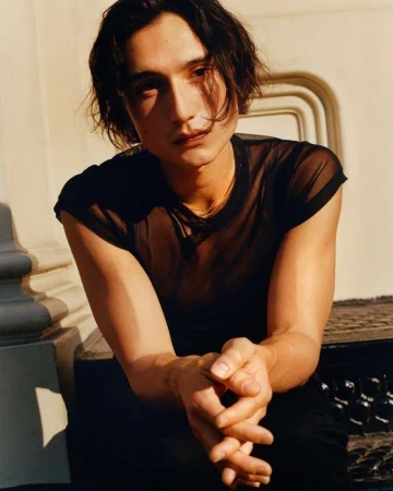 Portrait of a young adult male with dark hair and a sheer black shirt sitting outdoors against a light-colored concrete wall and steps.