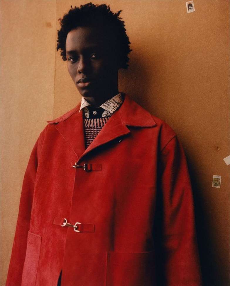 Portrait of a young Black man in red suede jacket, patterned shirt, dark hair, against a neutral background; fashion advertising and editorial.