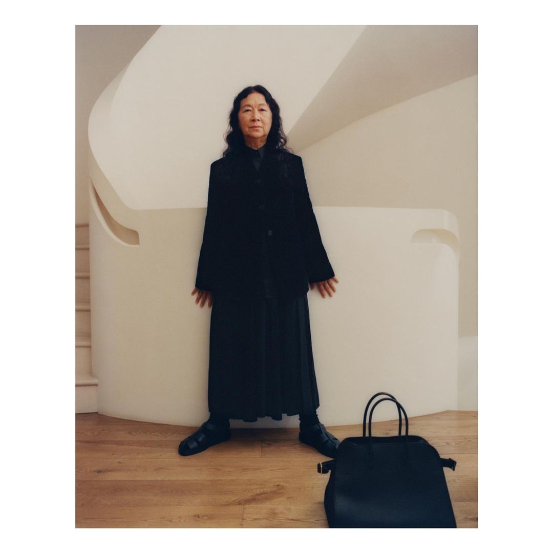 An Asian woman in a black coat and long skirt stands near a white curved staircase, complemented by a black purse on a wooden floor.