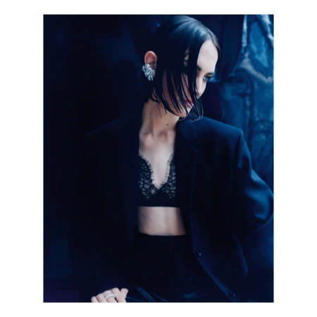 Portrait of a woman with dark hair wearing a black blazer, lace bralette, and statement ear cuff against a dark background