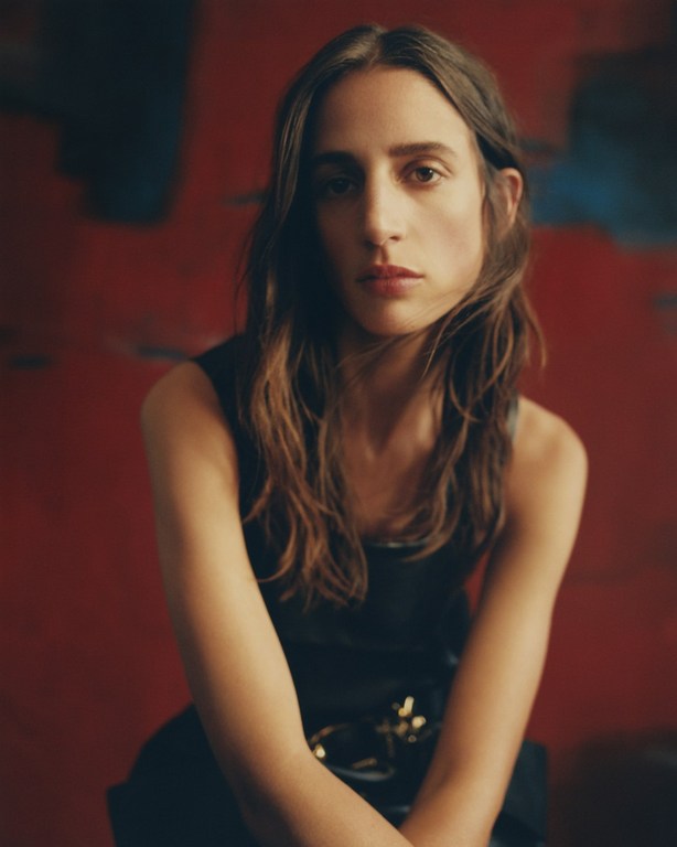 Close-up portrait of a woman with long brown hair, wearing a dark top and belt, against a red and blue painted background.
