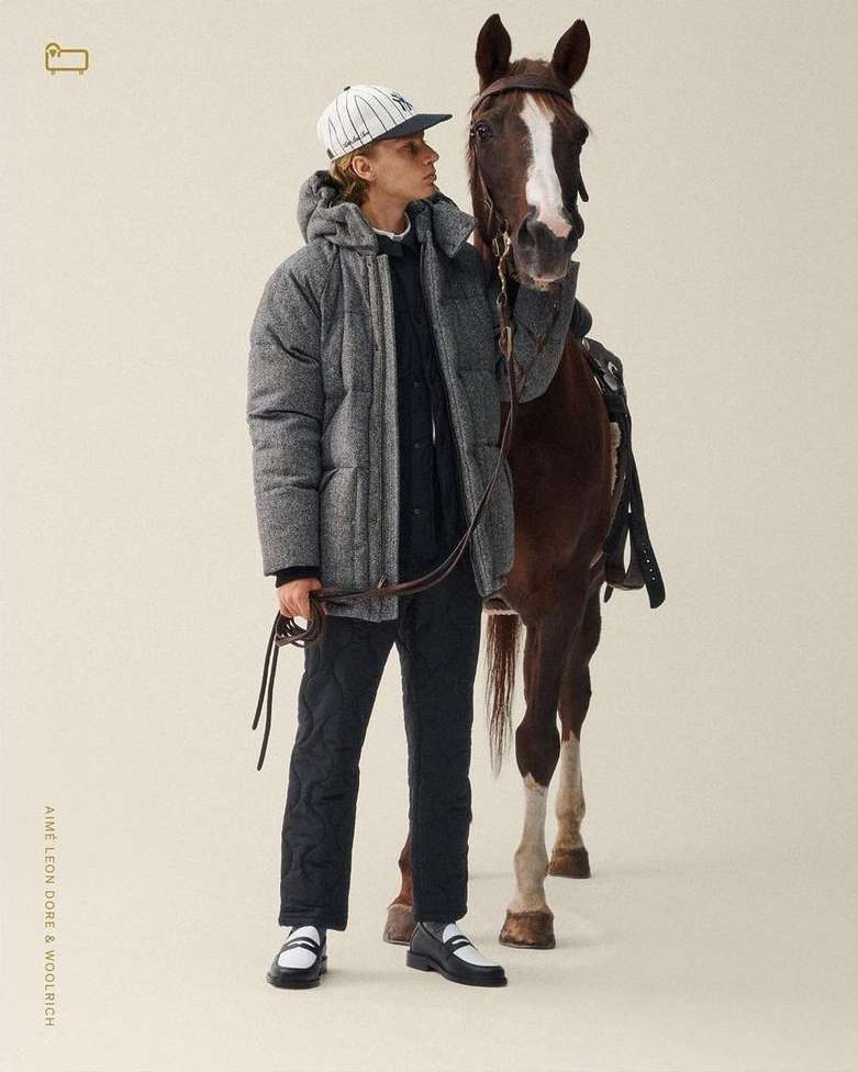 A young male model in a gray puffer jacket stands next to a brown horse in a neutral studio setting for an outdoor fashion shoot.