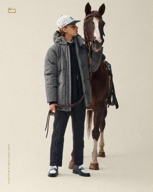 A young male model in a gray puffer jacket stands next to a brown horse in a neutral studio setting for an outdoor fashion shoot.