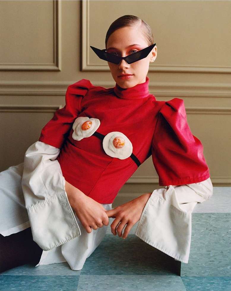 A person wearing a red top with egg-shaped accents and black sunglasses poses indoors against a paneled wall with a neutral backdrop.