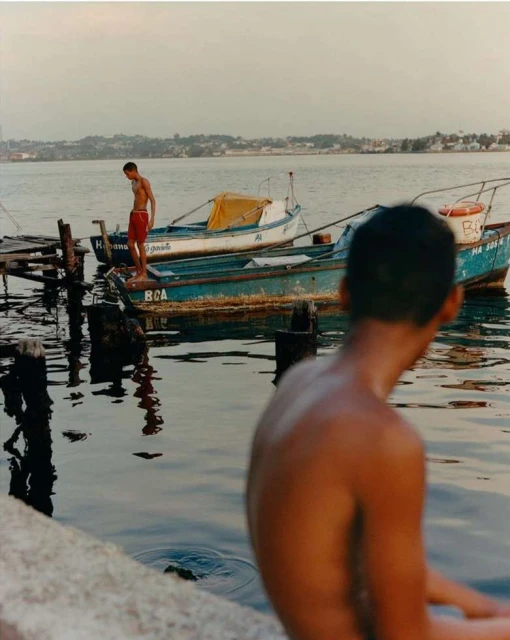 Two young men are by the sea with boats, one standing on a boat near a wooden pier, and another sitting by the water.