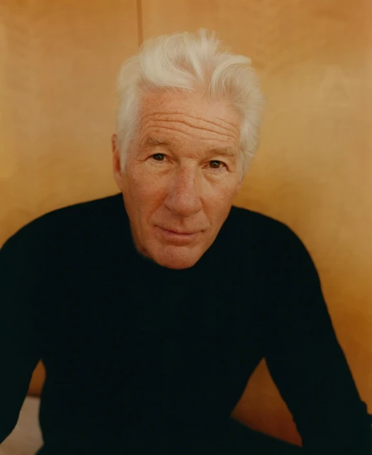 Headshot portrait of an adult male with white hair wearing a black sweater in front of a neutral brown background. Studio lit.