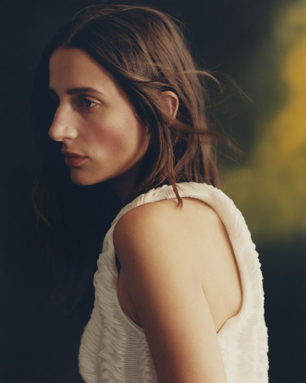 Portrait of a brunette woman in a textured white top, glancing over her shoulder against a blurred, dark green background.