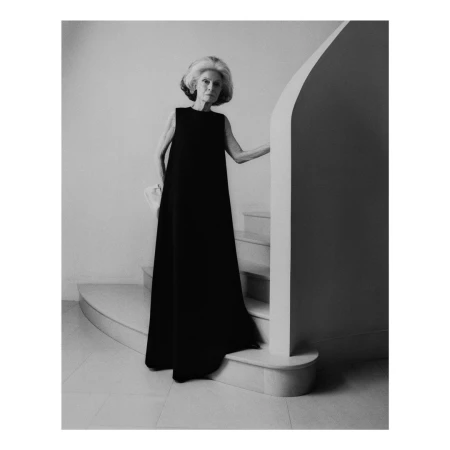 Elegant elderly woman in black sleeveless gown poses on stairs with white handbag, black-and-white studio photograph.