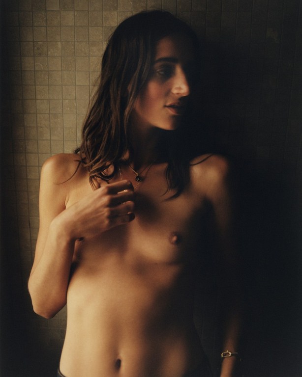 Portrait of a topless adult female with long dark hair, fair skin, and neutral expression posed indoors against a textured wall.