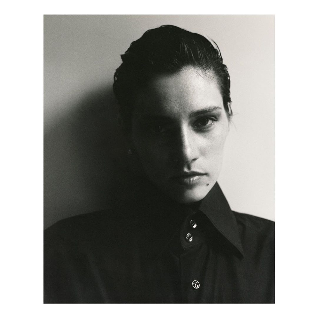 Portrait of a person with dark slicked-back hair wearing a dark collared shirt against a plain light wall.