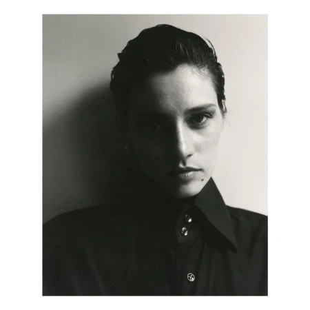 Portrait of a person with dark slicked-back hair wearing a dark collared shirt against a plain light wall.