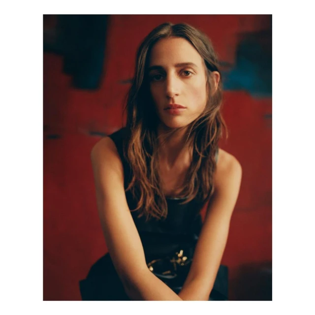 Portrait of a woman with long brown hair wearing a black sleeveless top, seated against a red abstract background.