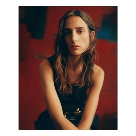 Portrait of a woman with long brown hair wearing a black sleeveless top, seated against a red abstract background.