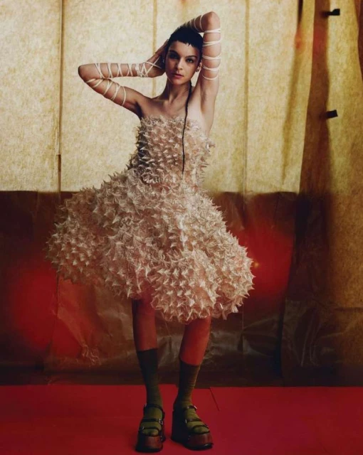 Fashion shot of a female model in an off-white textured dress, posing with hands behind her head against a textured gold backdrop.