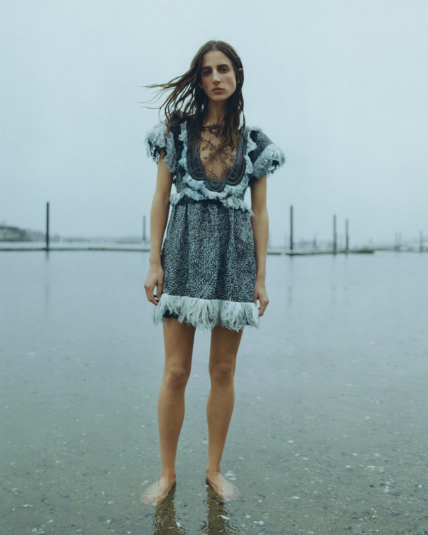 A woman in a patterned blue dress stands in water on a cloudy day, creating a fashion or editorial image with a reflective surface.