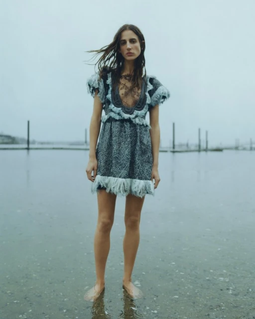 A woman in a patterned blue dress stands in water on a cloudy day, creating a fashion or editorial image with a reflective surface.