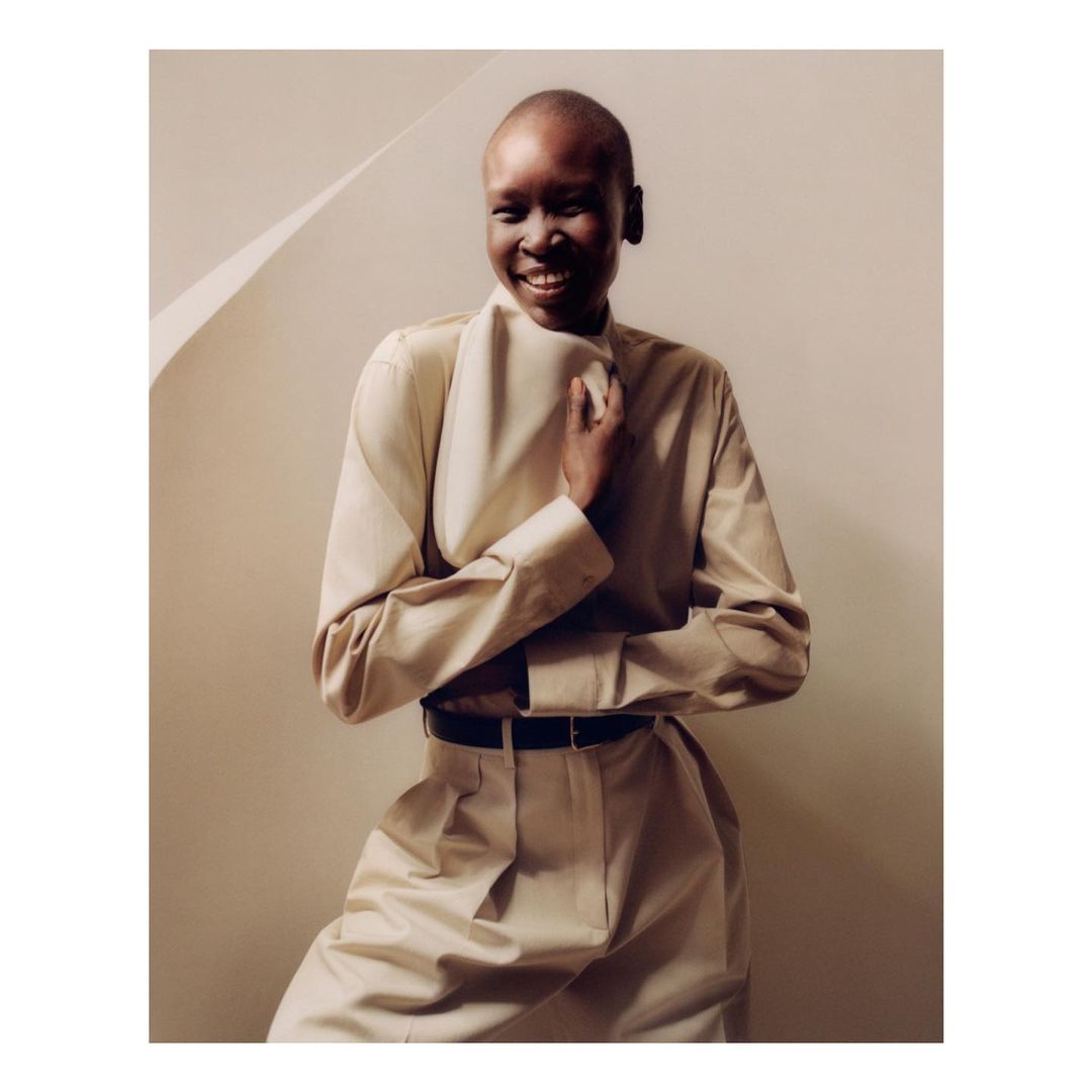 A smiling, bald, Black woman in a beige collared shirt and matching pants poses indoors against a beige wall and geometric shadow.